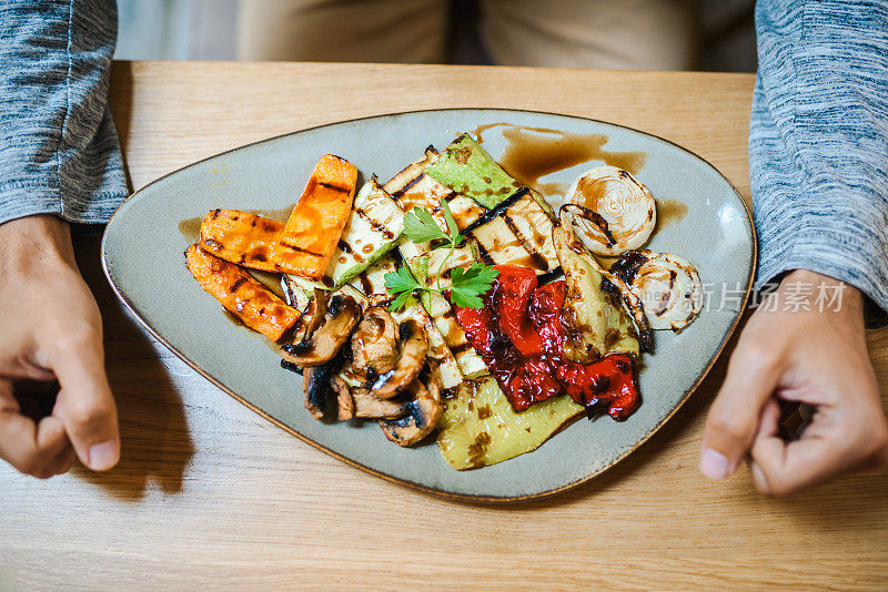 从上面拍下一个面目全非的男人的双手和一盘烤蔬菜。素食/餐厅里的素食菜肴。健康食品/饮食概念。